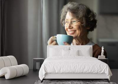 Happy senior woman drinking coffee looking out window at home, enjoying domestic rest, standing with cup in hands, looking aside and smiling Wall mural