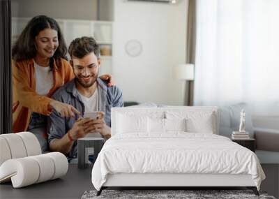 Happy young indian couple using smartphone, wife pointing at cellphone screen, sitting on sofa in living room interior at home. New app ad, banner Wall mural