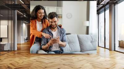 Happy young indian couple using smartphone, wife pointing at cellphone screen, sitting on sofa in living room interior at home. New app ad, banner Wall mural