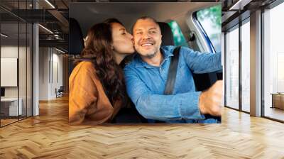 Loving woman kissing her smiling husband while driving car, sitting in auto and enjoying trip Wall mural