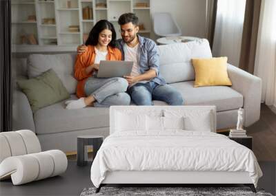 Planning vacation. Happy young indian couple searching tour online, using laptop computer, sitting on couch in cozy living room interior at home, free space Wall mural