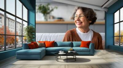 Portrait of happy senior woman relaxing on comfortable couch at home in living room and looking away at free space, enjoying domestic pastime Wall mural