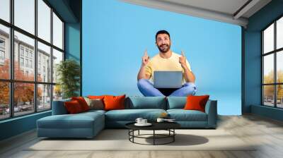 Portrait of happy young arabic man sitting isolated over blue background holding laptop on his knees and pointing fingers up Wall mural