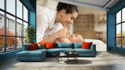 Smiling caring mother playing with cute little daughter in bedroom, mum and infant girl lying on cozy bed, holding hands, having fun, childcare concept Wall mural