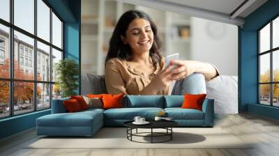 Smiling hindu lady using her smartphone while relaxing on couch at home, happy woman looking online content, sitting on grey sofa in living room, copy space Wall mural