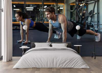 Young caucasian couple working out together, doing plank on dumbbells or push-ups on weights, training in modern gym interior Wall mural