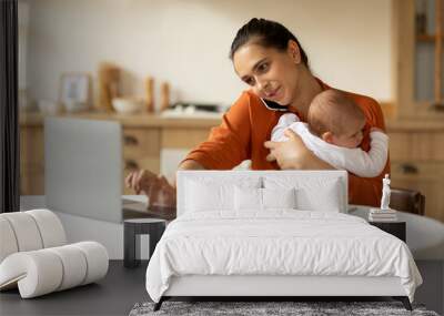 Young mother entrepreneur working from home while taking care of her infant little daughter, woman using laptop pc and talking on phone in kitchen Wall mural