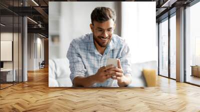 Young smiling hindu man holding mobile phone, typing sms or scrolling social media, sitting on couch in light living room interior, free space Wall mural