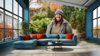 Young  cheerful curvy woman in grey hat and green jacket walking in the park Wall mural