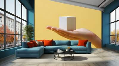 A hand holding a small white cube appears against a pale yellow background in minimalist composition, highlighting clean aesthetic and soft lighting for a professional photographic presentation. Wall mural