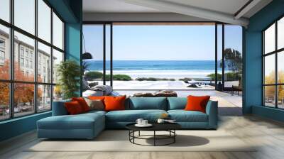 A large, sliding glass door with black frames on the left side of an open living room leading to beach in front of ocean, with blue sky and sand.
 Wall mural