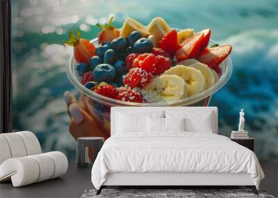 A person holding an acai bowl with strawberries, blueberries and bananas in their hand against the backdrop of sea water. This scene conveys freshness and natural beauty. Wall mural