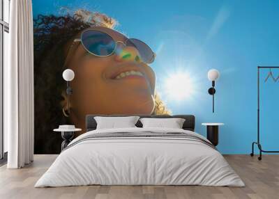 A woman wearing sunglasses smiles as she looks up at the sun on an sunny day, with blue sky in background. She is standing outdoors and has long curly hair. Wall mural