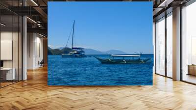 Catamaran and wooden boat in the sea off the coast of a tropical island. A catamaran with lowered sail and a wooden boat are anchored in the open sea with a tropical island in the background. Wall mural