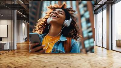 Joyful young woman immersed in music with wireless headphones, standing behind a towering building Wall mural