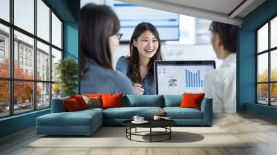 Three Japanese office workers are smiling and discussing work in the meeting room, with a laptop displaying financial data on the screen Wall mural