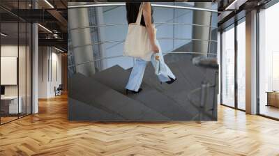 Stylish young woman wearing casual clothes and tote bag on shoulder walking down the stairs with a glass bottle of water in hand. Unrecognizable trendy female on walk in the city street Wall mural