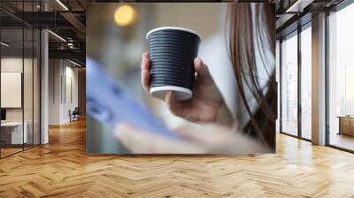 Young woman holding a disposable paper cup of coffee in hand. Female person drinking latte and browsing a social media app on a smart phone Wall mural