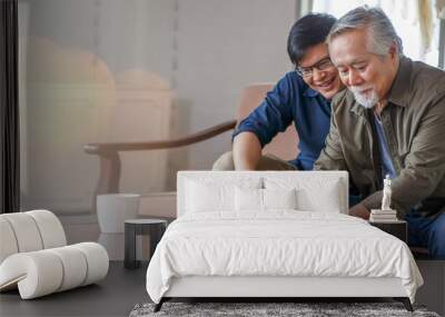 happy asian adult son and senior father sitting on sofa using laptop together at home . young man teaching old dad using internet online with computer on couch in living room . copy space banner Wall mural