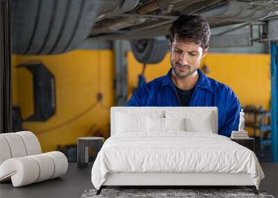 happy latin auto mechanic man checking tires , brakes under car with clipboard in garage cars service . hispanic technician repairing vehicle. lifted car checklist tyres and wheel in auto repair shop Wall mural