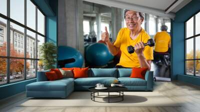 Senior asian sport man lifting dumbbells and show thumbs up in fitness gym . elder male exercising ,  working out , training weights, healthy ,Retirement , older, looking camera Wall mural