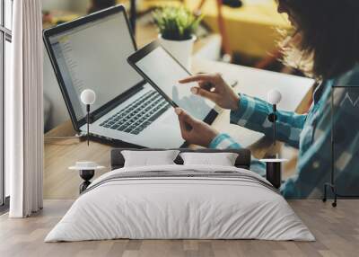 Close-up of young hipster girl sitting at workspace and using modern digital tablet and laptop, female businesswoman or designer working on devices at office, home interior on background Wall mural