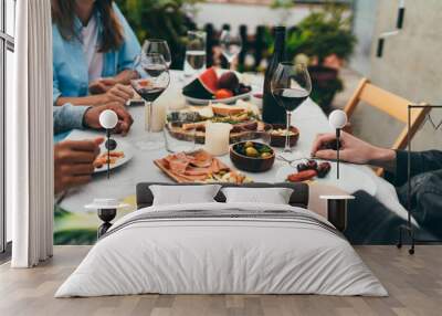 Group of young people dressed in casual clothes dining together drinking red wine and eating snacks, fruits, Friends celebrating birthday sitting in restaurant with small garden enjoying atmosphere Wall mural