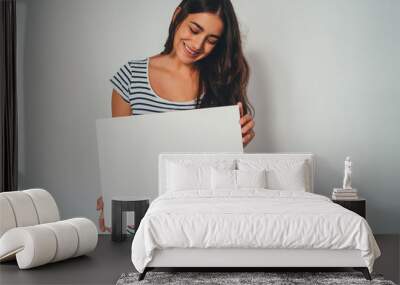 Happy young woman in casual t-shirt holding blank poster for your text message or design, Smiling Woman Holding Blank Paper Against White Background, Mock up of poster Wall mural