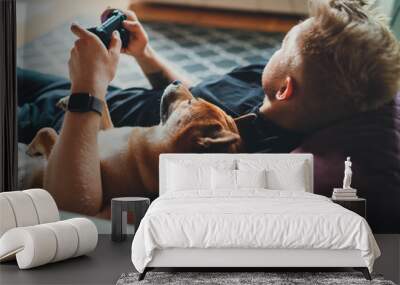 Young hipster guy playing video games at home holding game joystick relaxing on couch with his best friend dog, Home Leisure Concept Wall mural