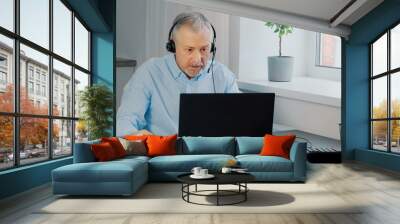 An elderly man wearing headphones sits at a laptop and makes notes. Wall mural