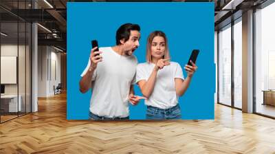 Brunet man in white tees and blue jeans is shocked looking at the smartphone screen of a blond woman. Wall mural