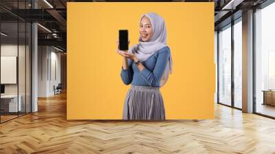 happy asian indonesian muslim woman holding smart phone and showing blank screen on isolated yellow background Wall mural