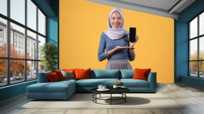 happy asian indonesian muslim woman holding smart phone and showing blank screen on isolated yellow background Wall mural