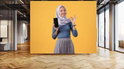 happy asian indonesian muslim woman holding smart phone and showing smart phone blank screen and giving ok finger gesture on isolated yellow background Wall mural