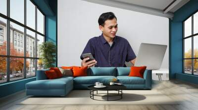 joyfull happy asian man holding mobile phone while sitting in front of laptop computer. on isolated background Wall mural