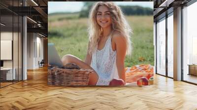 A cheerful young woman sitting on a picnic blanket in a sunny field, working on her laptop with a basket of fruits. Wall mural