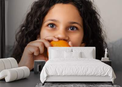 girl with tangerine. little tanned girl with dark long curly hair sits on a gray background and eats an orange tangerine, close-up fruit concept Wall mural