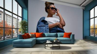 Confident lady in white t-shirt in jeans with denim jacket posing on camera in blue lighting holds hand on glasses. Fashion concept Wall mural