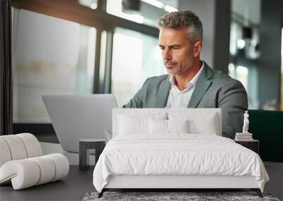 a busy, mature male executive, holding documents and working on a laptop, while attentively viewing his computer screen at his office desk Wall mural