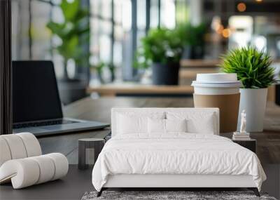 A laptop, notepad, and two coffee cups on a wooden table in a coffee shop with green plants in the background. Wall mural