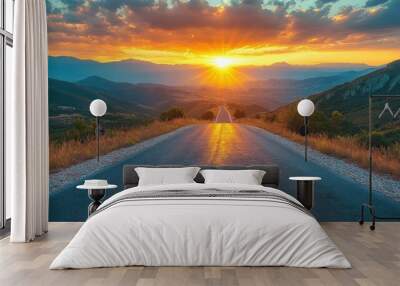 A winding asphalt road leads through a mountainous landscape towards a vibrant sunset. The sun's rays pierce through the clouds, casting a warm glow over the scene. Wall mural