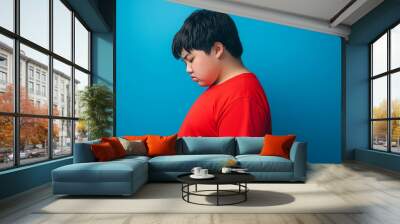 A side profile photograph of an overweight teen boy wearing a red t-shirt, standing with his head down, looking sad and dejected against a blue background Wall mural