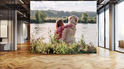 Child girl sitting and hugging golden retriever dog outdoors at the nature and looking at the lake. Teen kid with doggy pet resting close to river Wall mural