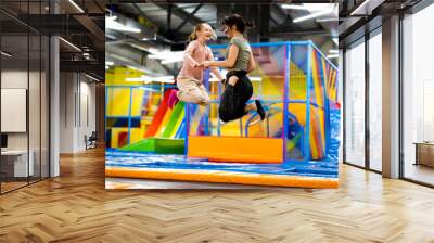 Pretty girls jumping together on colorful trampoline at playground park. Two sisters having fun during active entertaiments indoor Wall mural