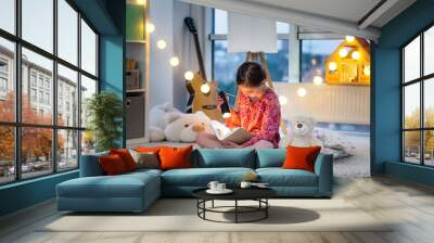 Pretty little girl with flashlight and book on floor in twilight room Wall mural