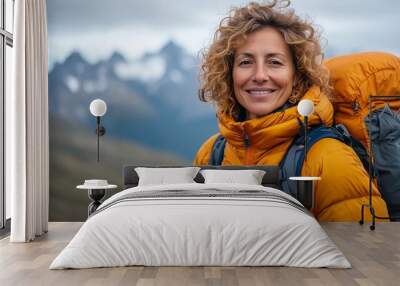 A middle-aged woman smiles at the camera while hiking in nature, wearing an orange down jacket and carrying backpacks on her back against the backdrop of mountains. Wall mural