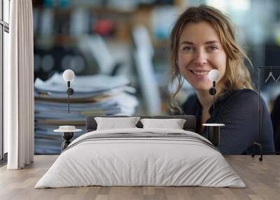 Female bookkeeper in office, sitting at desk with computer and pile of documents smiling to camera, Wall mural