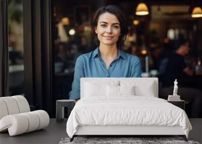 young female Barista waiter owner culinary gourmet bar restaurant worker in front of the shop with crossing arms wearing blue shirt, blurred shop in the background Wall mural