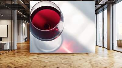   A close-up shot of a stemmed wine glass filled with dark red liquid resting on a plain white background, bathed in sunlight streaming through a nearby window Wall mural