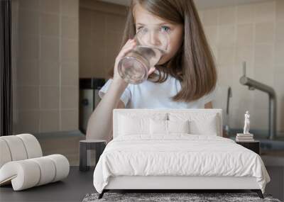 Happy adorable little girl drinking water in the kitchen at home. Caucasian kid  with long brown hair holding transparent glass in her hand. Indoors. Healthy life concept Wall mural